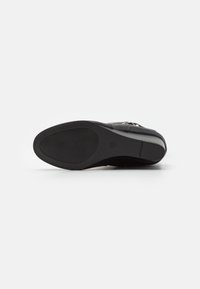 Semelle de chaussure noire avec caoutchouc noir lisse et motif de grip texturé ; présente un léger talon surélevé et un logo de marque en relief.