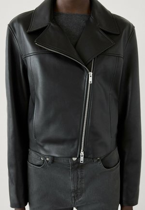 Personne portant une veste en cuir noire ajustée avec une fermeture éclair argentée asymétrique sur un pull gris foncé et un jean noir.