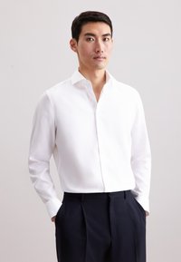 Camicia bianca strutturata con colletto aperto e maniche lunghe, abbinata a pantaloni blu scuro. La camicia presenta un motivo sottile.