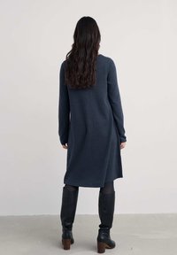 Nainen, jolla on pitkät, laineikkaat hiukset, pukeutuneena tummansinsiseen polvipituiseen mekkoon, mustiin sukkahousuihin ja mustiin korkokenkiin, seisoo yksinkertaista taustaa vasten.