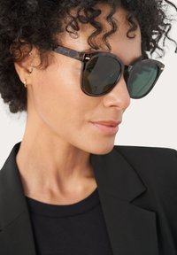 Occhiali da sole tartarugati con lenti scure, forma quadrata e dettagli dorati. Indossati con un blazer nero e una maglia, caratterizzati da una texture liscia.