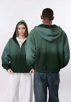 Nainen kasvot eteenpäin, yllään tumma vihreä vetoketjullinen huppari hupun kanssa, mies selin päin samassa hupparissa ja siniset farkut, yksivärinen tausta.