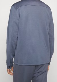 Suéter azul de manga larga, textura suave y diseño minimalista. Presenta costuras en los hombros y detalles de botones en los puños.