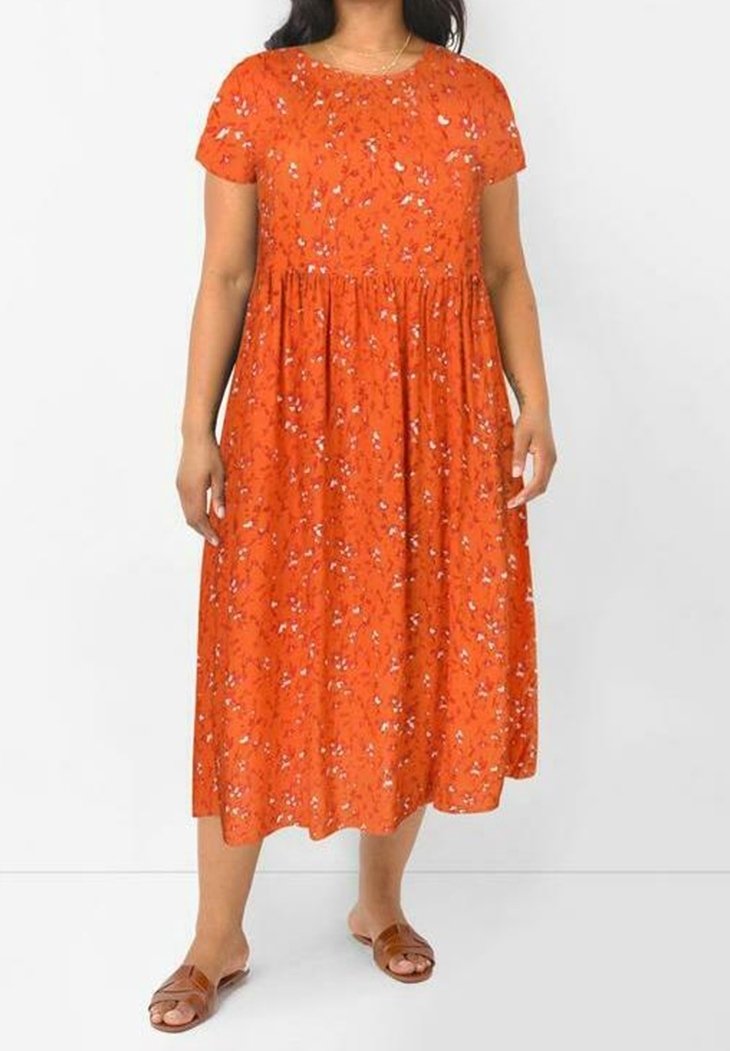 Nainen, jolla on lyhythihainen oranssi kukallinen midi-mekko ja ruskeat liukuvartiset sandaalit, seisoo yksinkertaisen valkoisen taustan edessä.