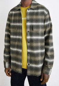 Grön- och vitrutig skjortjacka med tryckknappar och två bröstfickor, över en gul t-shirt, med mörkblå byxor.