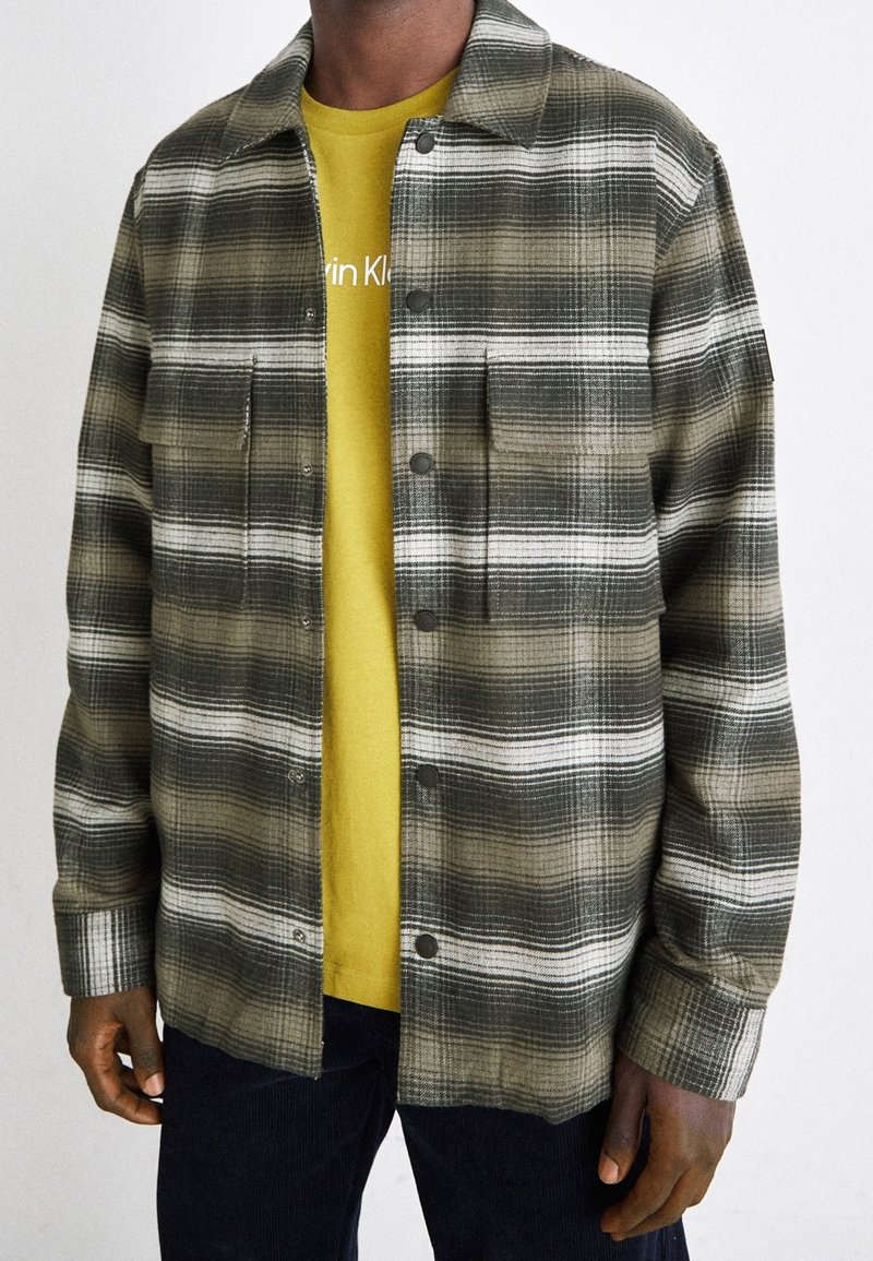 Grön- och vitrutig skjortjacka med tryckknappar och två bröstfickor, över en gul t-shirt, med mörkblå byxor.