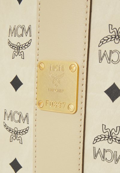Sac MCM avec une sangle en cuir beige, ornée d'une plaque en métal doré, d'un logo en relief et d'un fond crème avec un motif de diamants noirs.