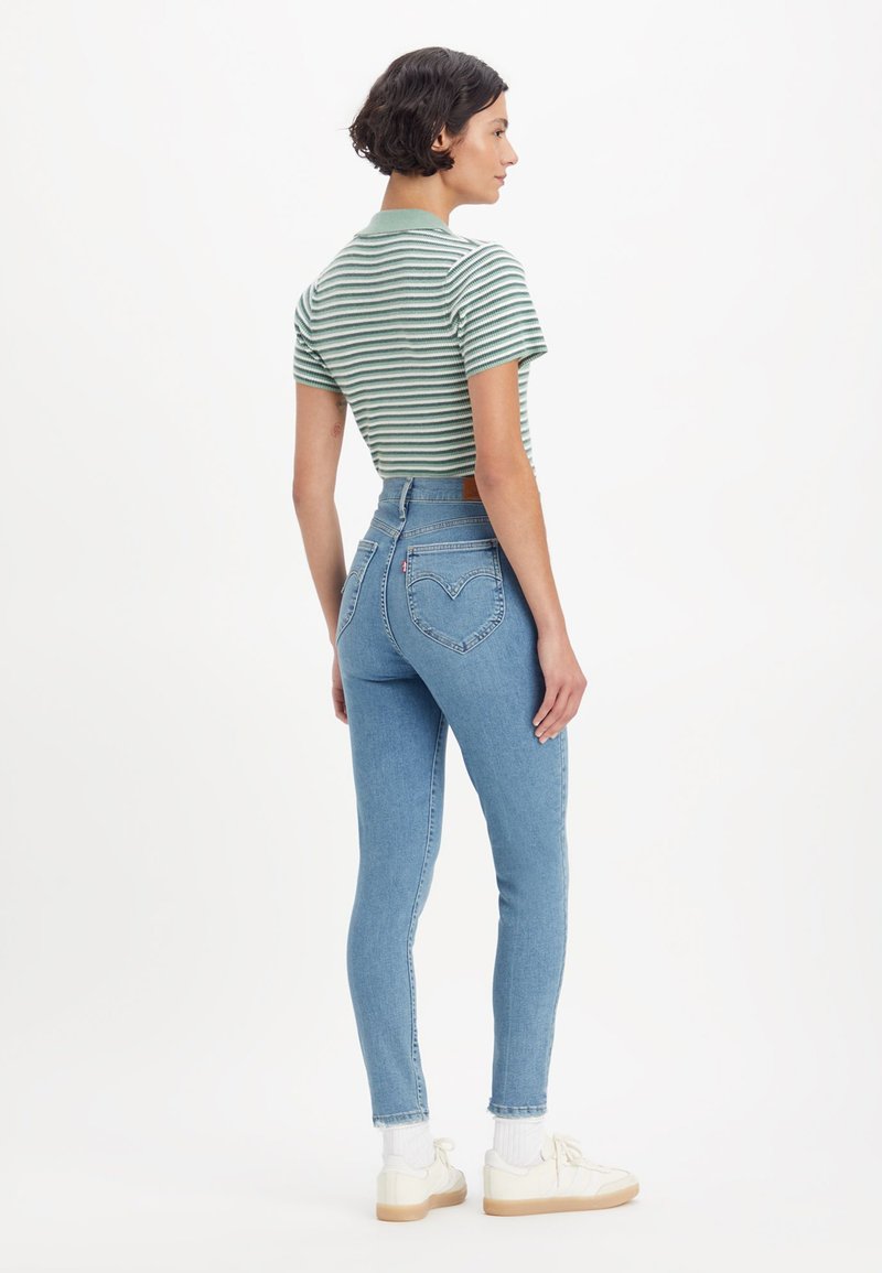Frau steht seitlich und trägt ein grün gestreiftes Poloshirt, hochgeschnittene hellblaue Skinny-Jeans mit herzförmigen Gesäßtaschen und weiße Sneaker.