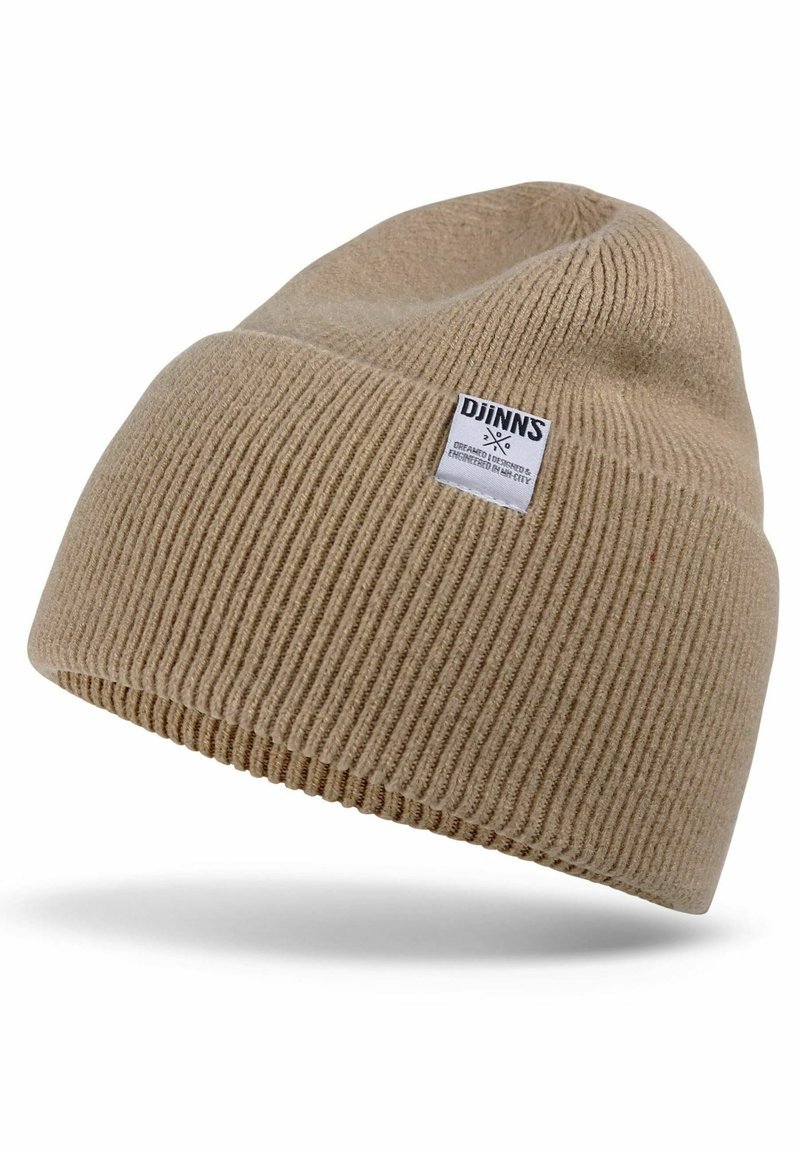 Beige knit beanie with ribbed texture. Features a small white label with black text. The design is soft and stretchy, suitable for cold weather.