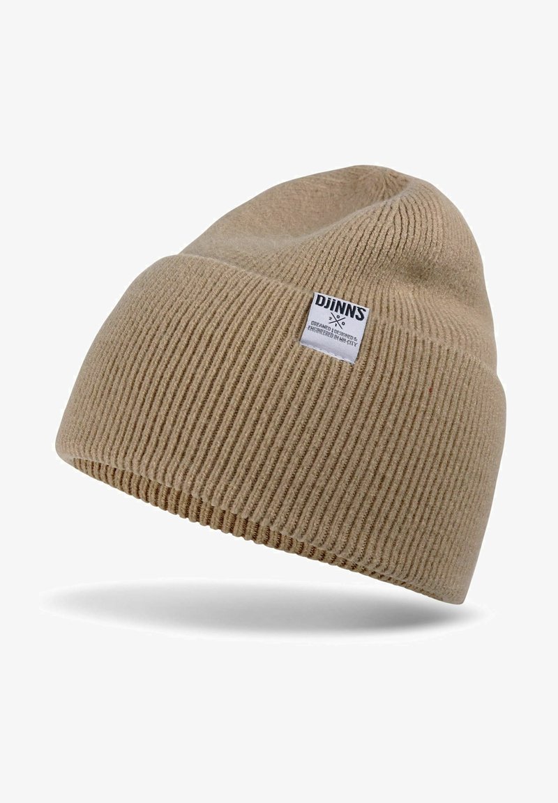 Beige knit beanie with ribbed texture. Features a small white label with black text. The design is soft and stretchy, suitable for cold weather.