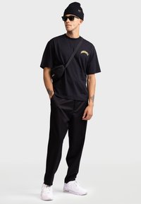 Svart bomullströja med en gul grafisk design, i kombination med svarta byxor och en svart kuvertväska. Vita sneakers och en svart mössa fulländar stilen.