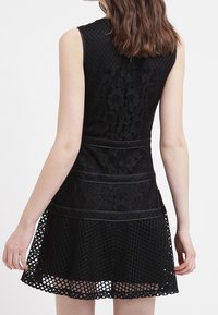 Svart ärmlös klänning med texturerad spets och en lager-på-lager-design, med en figursydd överdel, utsvängd kjol och meshinlägg vid kjolkanten.