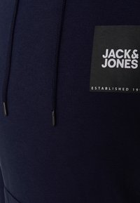 Námořnický modrý svetr má texturovanou tkaninu, nastavitelné černé šňůrky a bílou nášivku s logem "JACK & JONES" a rokem založení.