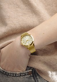 Reloj de metal dorado con correa de malla y una esfera de un dorado claro, con detalles texturizados y marcadores de hora. Lleva en una muñeca con un suéter beige.