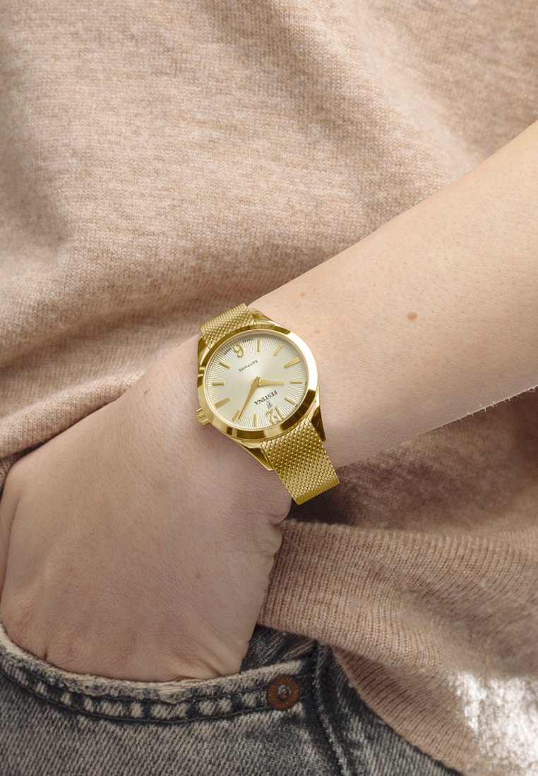 Reloj de metal dorado con correa de malla y una esfera de un dorado claro, con detalles texturizados y marcadores de hora. Lleva en una muñeca con un suéter beige.