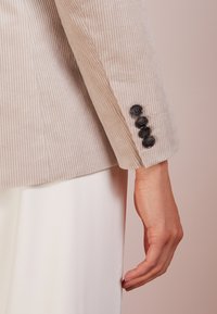 Beige Corduroy-Blazerärmel mit fünf schwarzen Knöpfen. Glatte Textur mit vertikalen Rippungen, taillierte Passform und elegantes Manschettendetail.