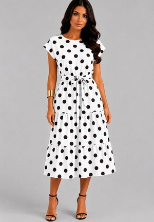 Femme portant une robe midi blanche à pois noirs, manches courtes, ceinture nouée à la taille et talons noirs à bride à la cheville sur fond gris.