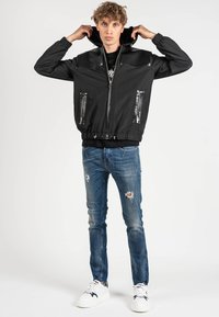 Giacca bomber nera con un collo in tessuto lucido, tasche frontali con zip, orlo elastico e cappuccio. Indossata su una maglietta nera e jeans strappati.