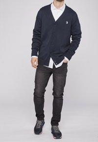 Mörkblå stickad cardigan med V-ringning, knäppning fram, texturerat tyg, bärs över en vit skjorta med krage, kombineras med mörka jeans och sneakers.