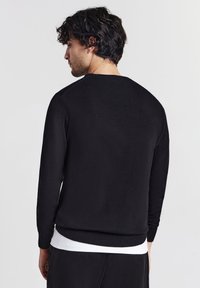 Uomo con i capelli neri e ricci, che indossa un maglione nero a maniche lunghe sopra una camicia bianca, rivolto di spalle su uno sfondo chiaro e uniforme.