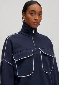 Marineblauwe ritscrede met hoge kraag, voorzien van twee voorzakken omlijnd in het wit. Zachte textuur met een relaxte pasvorm.