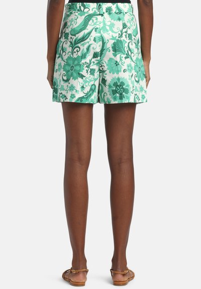 Femme portant un short blanc à motif floral vert et des sandales marron, vue de dos sur un fond clair uni.