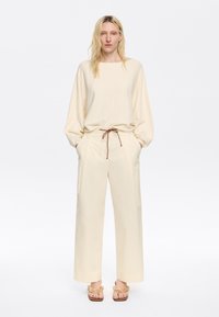Maglione lavorato a maglia beige con maniche ampie, abbinato a pantaloni larghi crema con coulisse marrone. Indossato con sandali beige a tracolla.