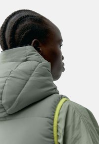 Padded grønn jakke med høy krage, med en teksturert finish. Jakken har et preg av lys gul langs stroppen.
