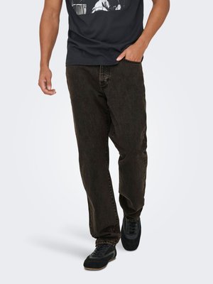 Homme marchant portant un t-shirt noir à motif, un jean marron foncé ample et des baskets noires sur un fond clair.