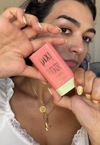 Il packaging di Pixi On-the-Glow Bronze è una bottiglia di colore corallo-rosa con scritte dorate e una base verde menta. Il prodotto è tenuto in mano con unghie dai toni neutri.