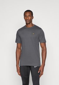 Pilkos spalvos medvilninė T-shirt marškinėliai su trumpomis rankovėmis, apvalia kaklo išpjova ir maža auksine logo ant krūtinės. Paprastas, kasdienis dizainas, lygus paviršius.