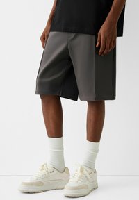 Shorts de sport gris en tissu lisse, avec une coupe ample et des détails cousus, associés à des chaussettes blanches mi-mollet et des baskets décontractées.