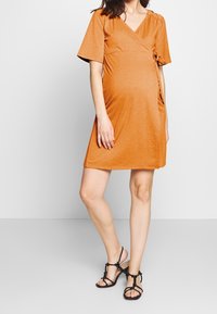 Pomarańczowa sukienka ciążowa wykonana z miękkiego materiału, z dekoltem kopertowym, krótkimi rękawami i detalem związywania z boku. Dobrana z czarnymi sandałami.