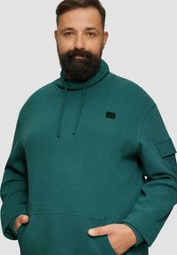 Sweatshirt teal avec un col haut, cordon de serrage, tissu texturé, poche avant et un petit logo noir sur la poitrine et la manche.