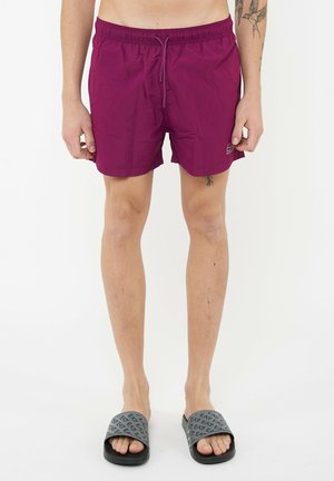 Hombre con pantalones cortos de baño color ciruela con cordón y sandalias grises tipo chancla con patrones de texto, de pie contra un fondo blanco liso.