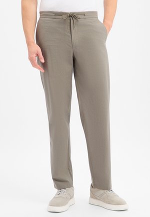 Homme portant un pantalon taupe droit avec taille à cordon et des baskets beiges, debout les mains dans les poches sur fond blanc.