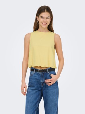 Ung kvinde smilende, iført gul ærmeløs top, blå højtaljede jeans, sort bælte og creoløreringe, stående mod en lys baggrund.