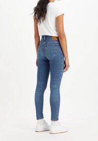 Strakke blauwe jeans van denim, met een hoge taille, achterzakken en een bruine leren patch aan de achterkant. Draagt een wit T-shirt en sneakers.