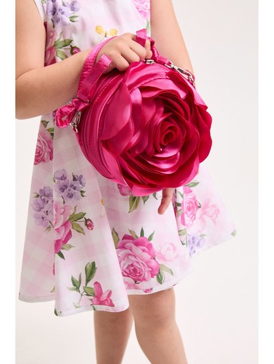 Νεαρό παιδί με φόρεμα με λουλούδια κρατά μια φωτεινή ροζ στρογγυλή τσάντα χειρός σε σχήμα τριαντάφυλλου, πάνω σε απλό φόντο.