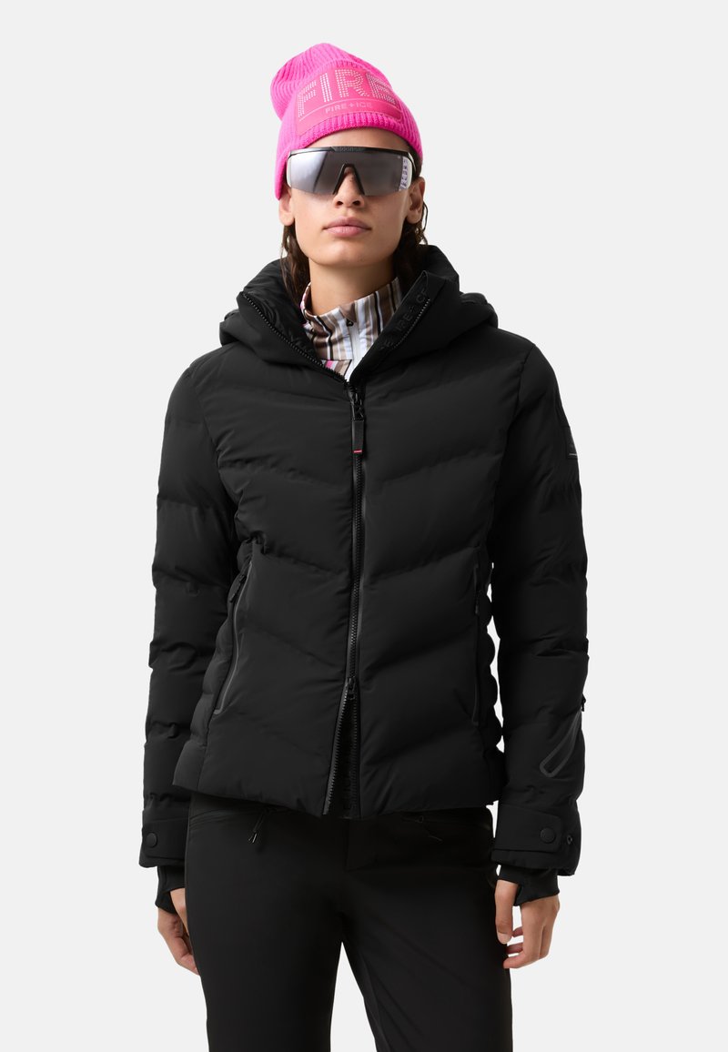 Sort pufferjakke med hætte, lynlåslukning og quiltet design. Bæret over en ternet skjorte, parret med en lys pink beanie og solbriller.