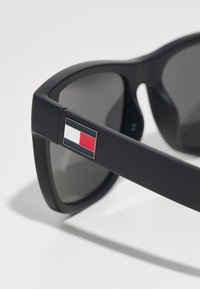 Gafas de sol negras con lentes oscuros, con montura rectangular. Detalle notable: acento del logo en el costado en colores rojo, blanco y azul.