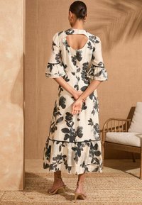 Cvetlična maksi obleka v kremni in črni barvi, z večslojnim robom, nabranimi rokavi in izrezom na hrbtu. Kombinirana z bež sandali z trakovi.