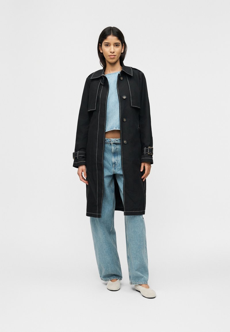 Γυναίκα που στέκεται φορώντας ανοιχτό μπλε τζιν παντελόνι και μπλούζα, κοντό μαύρο παλτό με κουμπιά και άσπρα επίπεδα παπούτσια, μπροστά σε απλό φόντο.