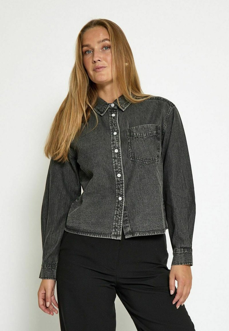 Donna con lunghi capelli biondi che indossa una camicia in denim nera sbiadita con bottoni e pantaloni neri, in piedi davanti a uno sfondo chiaro e uniforme.