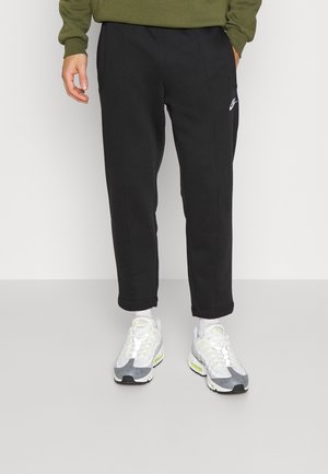 Person trägt schwarze Nike-Sweatpants, weiße und graue Nike-Sneaker und einen olivgrünen Pullover, steht vor einem schlichten Hintergrund.