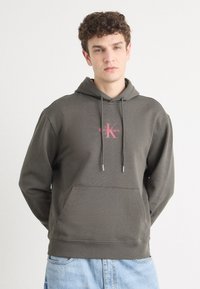 Hoodie verde oliva scuro in morbido tessuto con una tasca frontale, cappuccio regolabile con cordino e logo rosa di Calvin Klein sul petto.