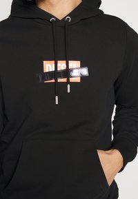 Sudadera negra con capucha y bolsillo frontal, con un logo rectangular rojo que dice "DIESEL" y una banda texturizada negra a lo largo. Ojales plateados y cordones.