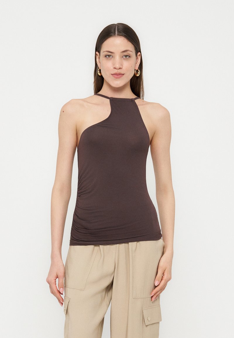 Γυναίκα με ίσια σκούρα μαλλιά φοράει αμάνικο ασύμμετρο καφέ τοπ και μπεζ παντελόνια τύπου cargo, στέκεται μπροστά σε λευκό φόντο.