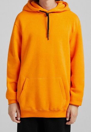 Personne portant un sweat à capuche orange vif avec un cordon noir et une poche kangourou avant, debout devant un fond uni.