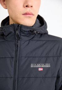 Mand iført en sort lynlåsjakke med hætte, der har Napapijri-logoet og et lille norsk flag på brystet.